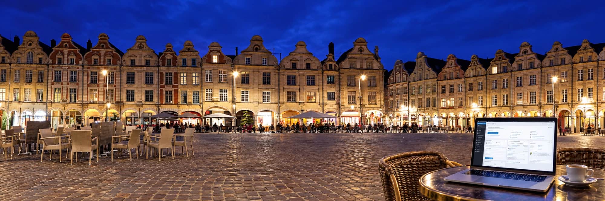 La Place des Héros d'Arras à l'heure bleue, ses façades baroques éclairées, et sur une table de terrasse un ordinateur portable dont l'écran affiche le tableau de bord de WordPress, à côté d'un café